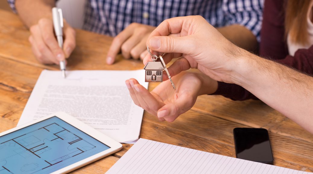 Happy young couple getting keys of their new house. Close up of female hand receiving keys from house broker. Closeup of the hand of a real estate agent who give the house keys to a woman while her boyfriend signing a contract.