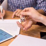 Happy young couple getting keys of their new house. Close up of female hand receiving keys from house broker. Closeup of the hand of a real estate agent who give the house keys to a woman while her boyfriend signing a contract.