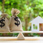 Home loan / reverse mortgage or transforming assets into cash concept : House paper model , US dollar hessian bags on a wood balance scale, depicts a homeowner or a borrower turns properties into cash