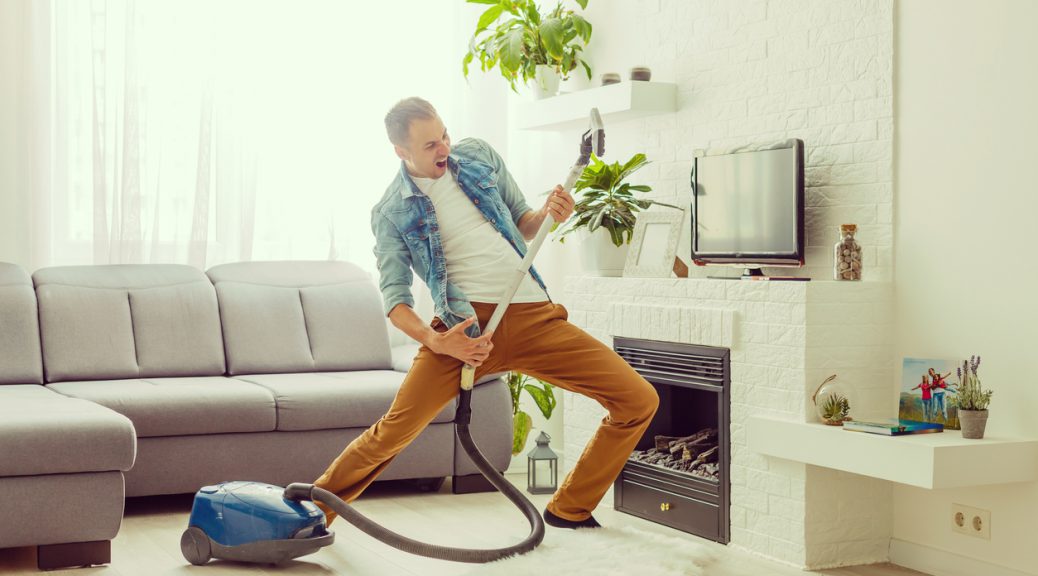 Young man having fun cleaning house with vacuum cleaner dancing like guitarist