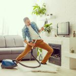 Young man having fun cleaning house with vacuum cleaner dancing like guitarist