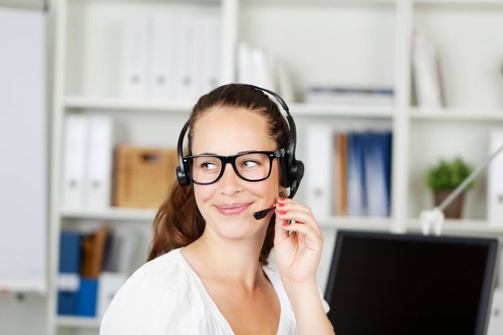 Smiling office worker wearing glasses and a a headset answering calls for a call centre or client services help desk
